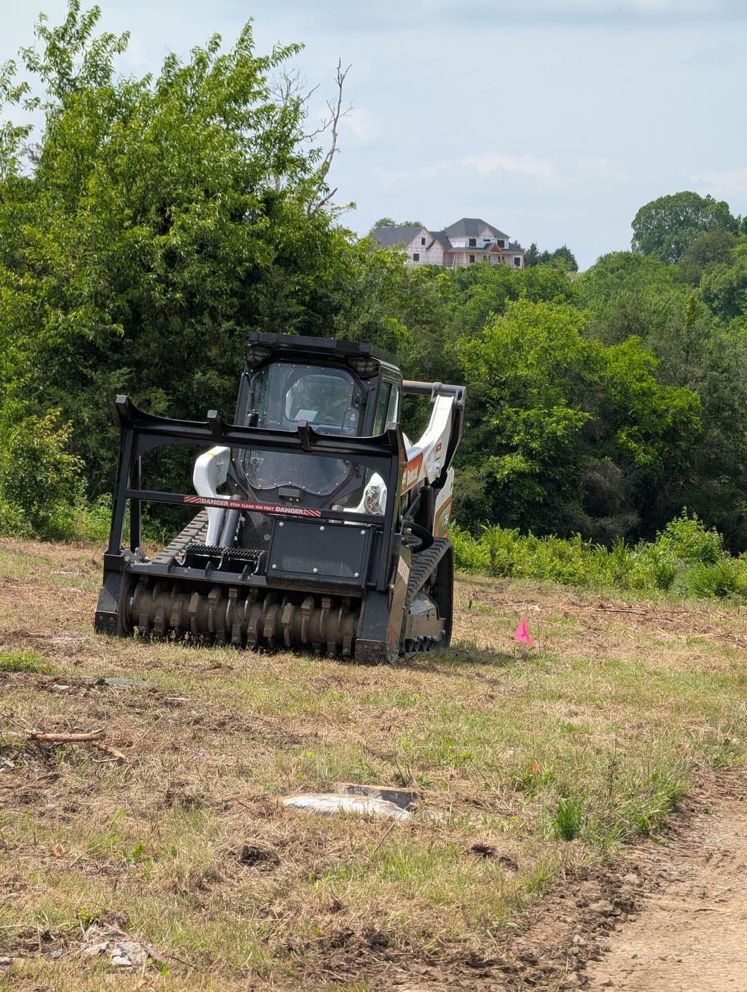 Bobcat skid steer with tiller attachment clearing land near a wooded area and a house.