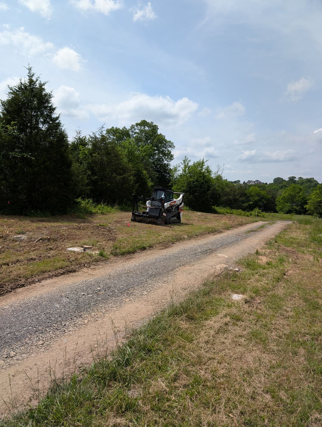 Excavator working on a gravel road in a wooded area under a blue sky.